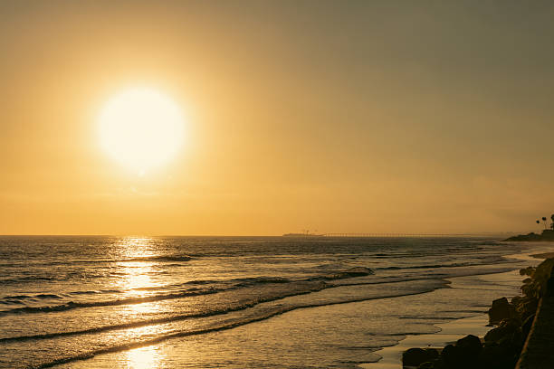 A wide view of Rincon Beach where a golden sunbeam shining through red and dark clouds reflects on the sea, creating a beautiful landscape. Gentle waves are breaking on the shore.