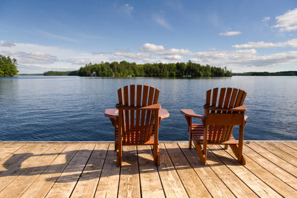 istock Adirondack chairs on a dock, facing a lake and a cottage view. 2205004343