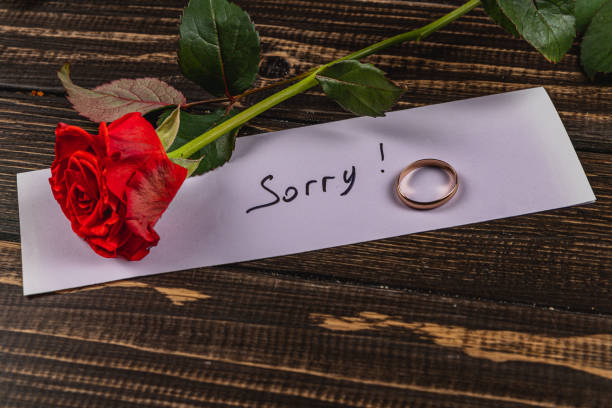 A red rose lies beside a wedding ring and a note that reads Sorry on a rustic wooden table, symbolizing the end of an engagement and conveying deep emotion.