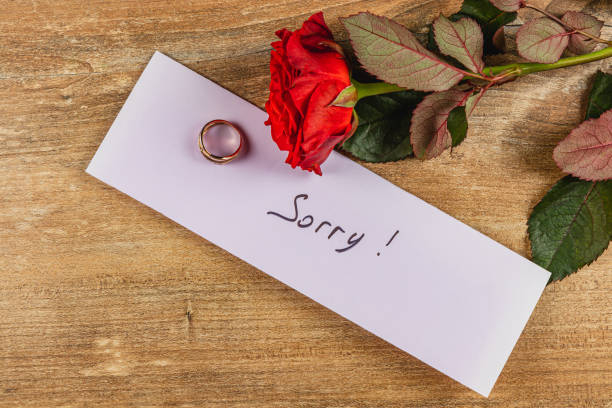 A broken engagement is expressed through a handwritten note saying Sorry, accompanied by a rose and a ring on a wooden surface, capturing the essence of lost love and regret.