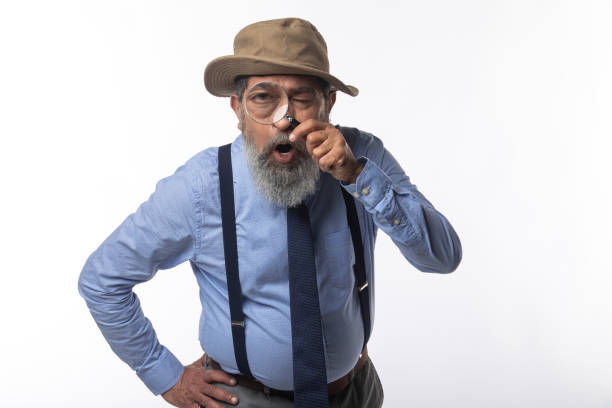 Portrait of senior detective wearing hat and suspenders looking through magnifying glass while standing on white background