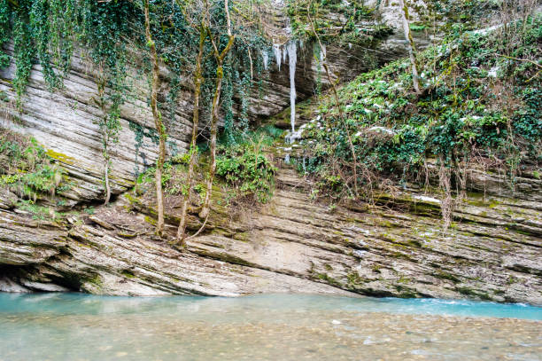 Clear blue water flows gently past moss-covered rocks and lush greenery, surrounded by steep cliffs in a peaceful natural setting.