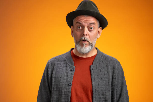 A studio portrait of a mature Caucasian man, wearing a gray cardigan and hat, displaying a surprised expression, set against a vivid orange background.