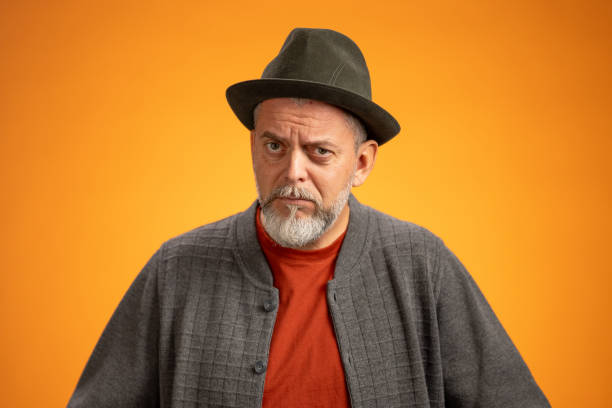 Studio portrait featuring a mid-aged Caucasian man with grey beard, wearing a fedora hat and textured grey jacket over rust-colored shirt, photographed against seamless orange backdrop.
