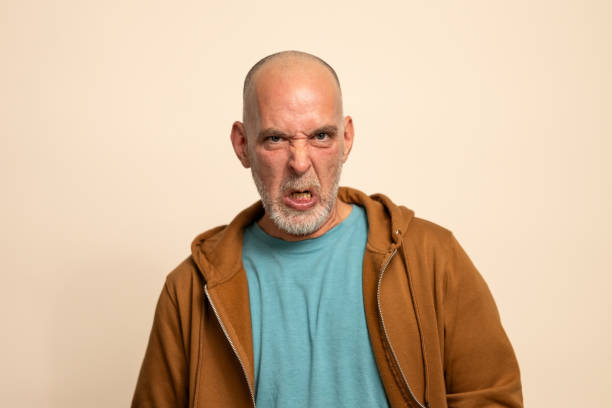 Studio portrait of mid-aged Caucasian male with bald head and grey beard displaying intense angry expression. Subject wears brown zip-up hoodie over turquoise t-shirt against neutral backdrop.