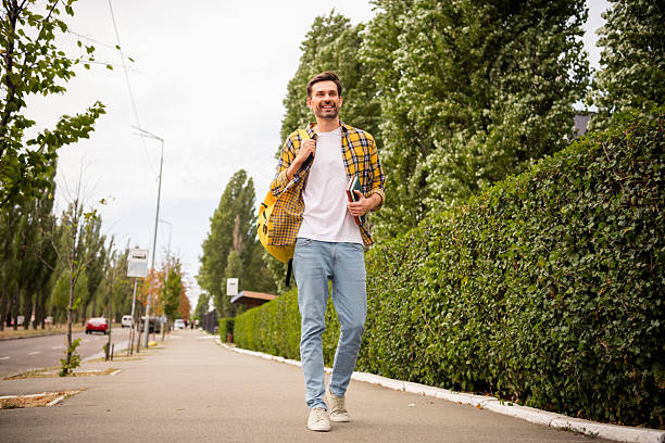 istock Young fashionable man walking outdoors during daytime with a cheerful expression, carrying books and a backpack 2205035088