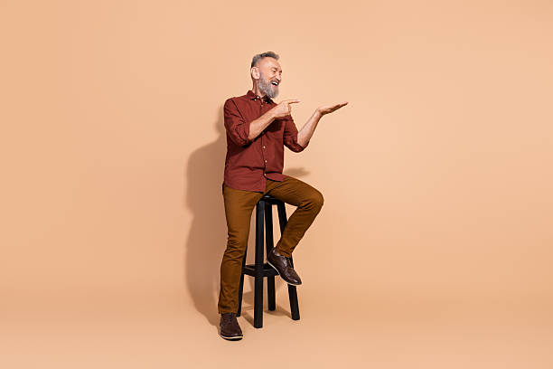 istock Mature man with grey beard and casual elegance posing on stool against beige background, gesturing confidently for fashion 2205039254