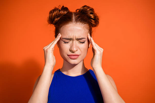 Young woman with a pensive expression touching her temples against a bright orange background, wearing a stylish blue top