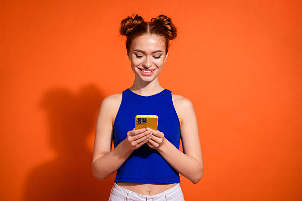 istock Young Stylish Woman with Red Hair Smiling While Using a Smartphone Against Bright Orange Background 2205039924