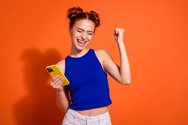 Excited Young Woman with Stylish Hairstyle Celebrating Success Holding Smartphone Against Vibrant Orange Background stock photo