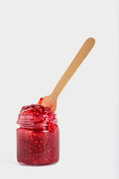 Glass jar with overflowing raspberry jam and a wooden spoon inside the preserve, white background.