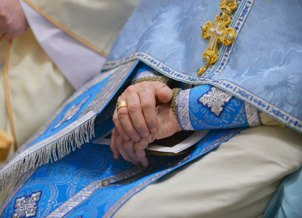 A close-up shot capturing the dignified presence of a bishop, his hands gently folded over his embroidered blue and gold vestment. A beautifully crafted cross pendant hangs from his neck, symbolizing his devotion and leadership in the Christian faith. The intricate gold details on his robe and the delicate texture of the fabric highlight the richness of religious tradition. The softly blurred background enhances the depth, making the focus entirely on the bishop’s sacred attire and spiritual presence.