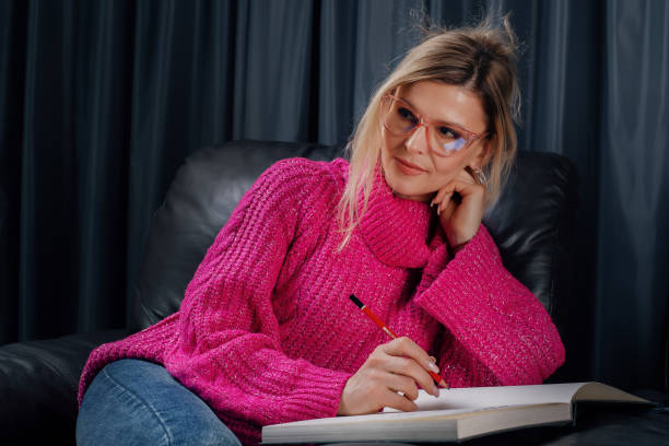 istock Young woman wearing pink sweater and glasses is writing on a notebook while sitting on a armchair 2205062410