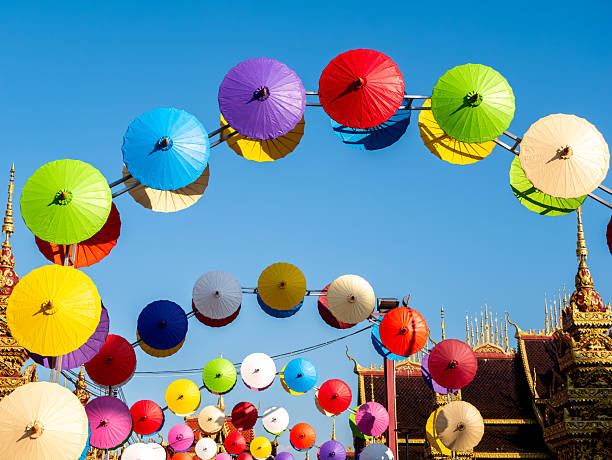 A vibrant display of colorful umbrellas hangs overhead at Wat Phiphat Mongkhon in Sukhothai, Thailand. The umbrellas create a festive canopy over a street within the temple grounds, with the beautiful temple architecture visible in the background. This image captures the joyful atmosphere and cultural richness of Thai temple celebrations A vibrant display of colorful umbrellas hangs above the golden gates of Wat Phiphat Mongkhon in Thung Saliam, Sukhothai, Thailand. The umbrellas create a cheerful and welcoming atmosphere, contrasting beautifully with the ornate golden architecture of the temple entrance