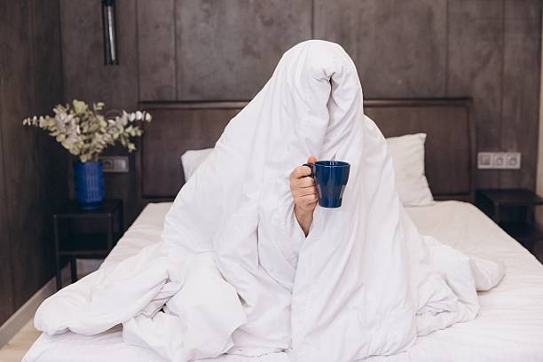 istock Sleepy Man Hiding Under Blanket Holding Coffee Cup in Bed 2205071531