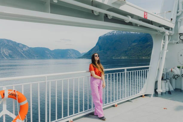 Side view of of happy female with long hair in orange t-shirt and pink pants enjoying the trip by the ferry through majestic fjord with scenic view of sea and the mountains in Western Norway, Scandinavia