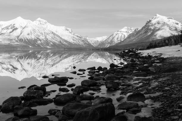 Glacier National Park Centered Mountain Range Showing Mirrored Reflection in Water/Lake with Snow/Ice In Foreground on a bed of stones on the shore - Clouds in the Sky - Black and White Photo