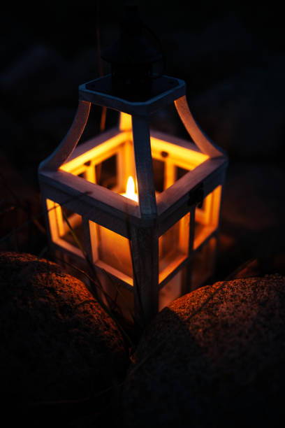 Close-up view of a rustic wooden lantern with glass panes, housing a flickering candle, nestled among rugged rocks in a dark nighttime setting. The soft, warm glow of the candle contrasts with the cool darkness, creating an intimate and atmospheric composition. This image conveys a sense of tranquility, solitude, and warmth, making it ideal for themes related to mindfulness, meditation, spirituality, and rustic aesthetics.