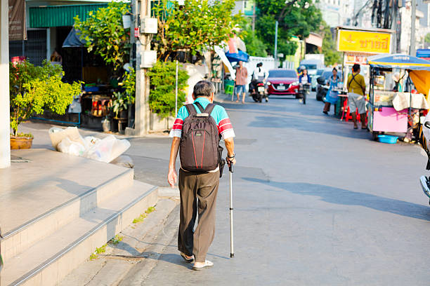 Rear view of walking thai senior man with cane. Man is walking parallel to Ratchadaphisek Road in Bangkok Chatuchak. Between road and sidewalk is this small street called Chaloem Suk Alley. Man is passing along a Seven-Eleven shop. He has a backpack for laptop branded Acer.  In background are some street food kitchens and another senior man with cane is walking there.