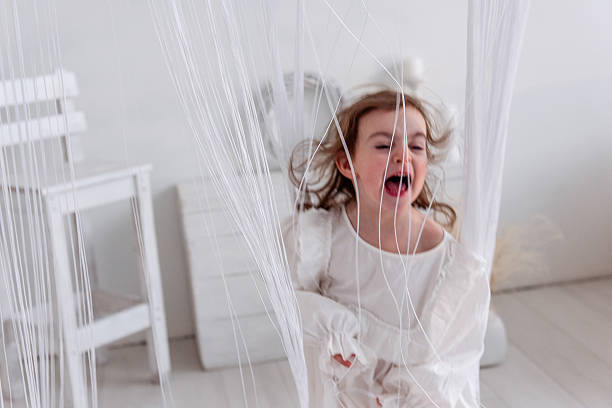 Excited preschool girl in a white dress running and shouting joyfully behind sheer white curtains, fully immersed in playtime within a bright, cheerful room filled with white furniture