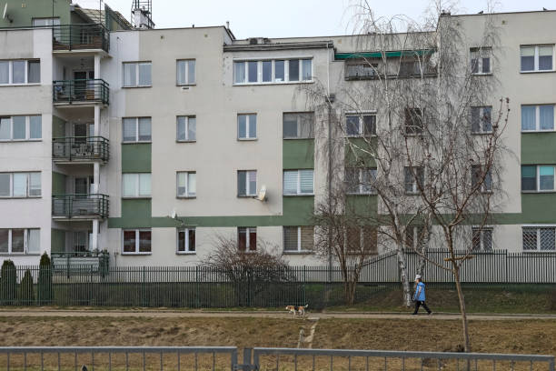 Warsaw, Poland - March 9, 2025: View of the Goclaw housing estate. The apartment building behind a fence, along which there is a path and a woman walking there. A birch growing beside the building.
