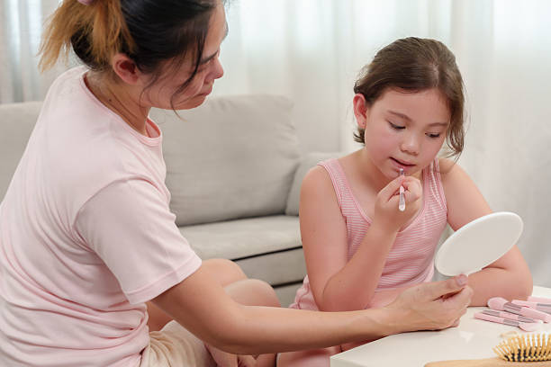 istock Asian mother holding mirror while Caucasian daughter applies lipstick, learning makeup techniques. Skincare products, makeup brushes, beauty items neatly arranged on white table in cozy living space. 2205106052