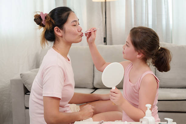 Caucasian daughter applying lipstick on Asian mother's lips while holding handheld mirror. Beauty products, skincare items, and makeup brushes neatly arranged on white table in bright home setting.