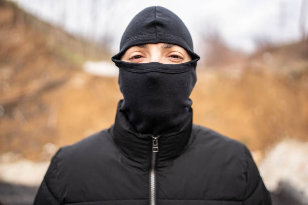 A focused portrait of an individual wearing a black mask and jacket outdoors in natural light. The background contains earthy tones, providing contrast with the dark clothing.