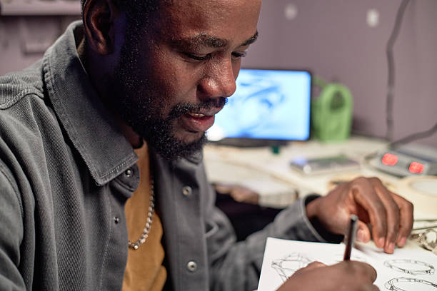 Man focusing while drawing detailed sketch on paper, a computer with design software in background. Engaged in creative work environment without distraction