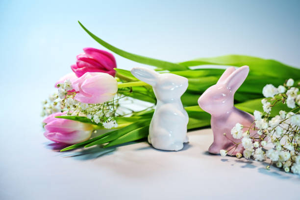 Two Easter bunnies from ceramic with a bouquet of pink tulips against a light blue background, holiday greeting card, copy space, selected focus, narrow depth of field