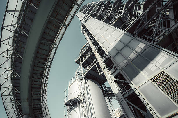 A towering oil refinery structure with industrial metal frameworks, storage tanks, and pipelines. Captured from a low-angle perspective, showcasing engineering complexity