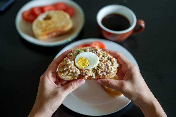 A close-up of hands holding a slice of toasted bread topped with tuna salad and a boiled egg, served with coffee and sliced tomatoes in the background, creating a cozy breakfast setting.