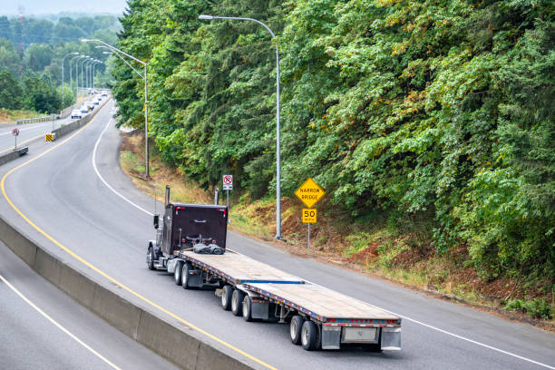 istock Black classic powerful big rig semi truck tractor transporting two empty flat bed semi trailers running on the divided highway road with green forest on the side 2205184979