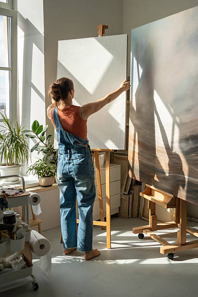 istock Artistic female holding canvas in hands standing in bright sunny workshop, preparing studio for work 2205189553