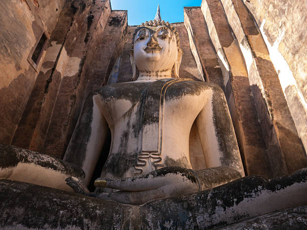 An ant's eye view of a large stucco Buddha image, nearly full figure, known as Phra Achana, within Wat Si Chum in Sukhothai Historical Park. Surrounded by weathered ancient brick walls and a bright blue sky, the image conveys the grandeur, sanctity, and age of this historic siteA nearly 800-year-old stucco Buddha image serenely smiles through the ancient brick walls of Wat Si Chum in Sukhothai, Thailand. The weathered brick and crumbling pillars in the foreground add to the sense of history and timelessness. This iconic image represents the rich cultural heritage and artistic legacy of the Sukhothai Kingdom