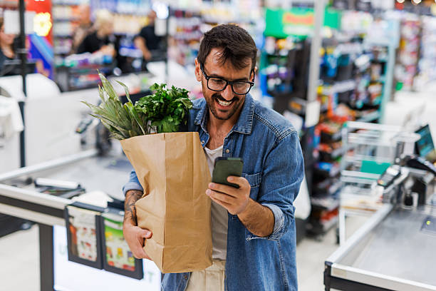 istock Smiling customer using smartphone while holding grocery bag in supermarket checkout line 2205225252