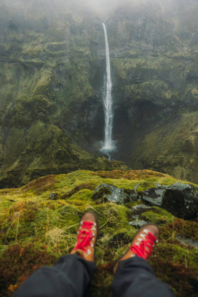 Personal perspective of women's legs wearing red hiking boots sitting at the edge of cliff on soft green moss, looking at ,majestic waterfall in canyon covered by fog in Vatnajokull National park, South Iceland