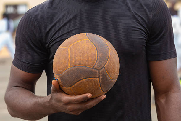 close-up shot of a person holding an old, worn-out leather football. The individual is wearing a black athletic t-shirt, and the background is slightly blurred, suggesting an outdoor sports environment. The image highlights the texture and history of the vintage ball, evoking themes of tradition, endurance, and the timeless love for the game
