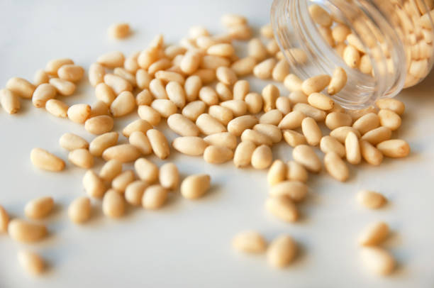 Pine nuts spread over white background, mouth of jar on the right, angle view