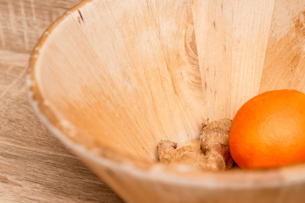 Close-up of a natural wooden bowl containing a fresh orange and ginger root, set against a wooden background. The minimalistic composition emphasizes organic textures and warm earthy tones, evoking a sense of natural living and healthy eating. This image is ideal for illustrating concepts related to nutrition, organic food, rustic kitchen decor, and wellness.