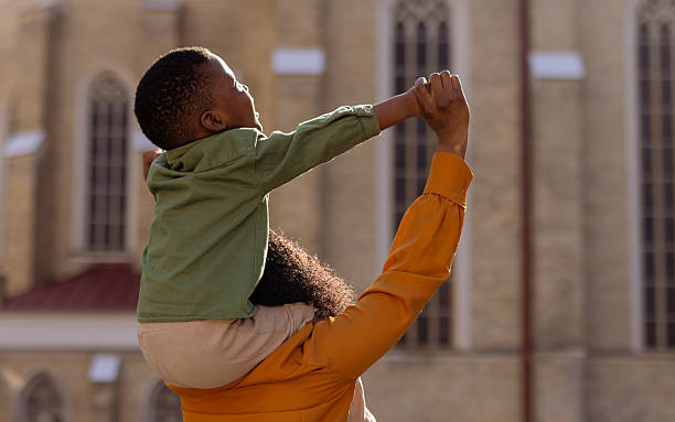 A black mother and son share a heartwarming moment while boy sitting on her mom's shoulders, enjoying a fun-filled day in the city