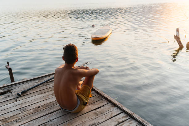 Teenage boy is sitting on the dock with his feet near the water, watching his paddle board in the lake. The lake is calm, and the background is filled with green trees and hills, creating a peaceful and relaxing atmosphere.