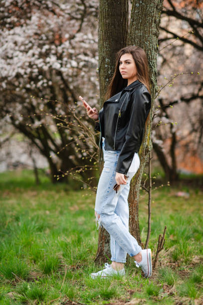 A young woman with long brown hair leans against a tree while looking at her smartphone, surrounded by blooming cherry blossoms. She wears a black leather jacket, ripped jeans, and white sneakers, blending urban fashion with a natural spring setting. The soft lighting and greenery create a fresh and modern atmosphere.