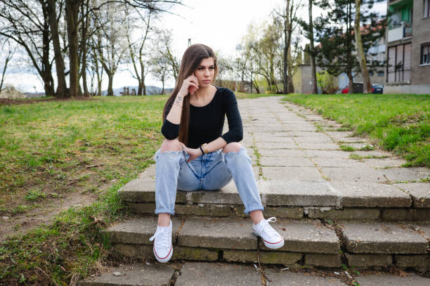 A young woman with long brown hair sits on outdoor concrete steps, resting her chin on her hand with a thoughtful expression. She wears a black long-sleeve top, ripped jeans, and white sneakers, combining a modern and casual style. The natural surroundings and soft lighting create a calm and introspective atmosphere.