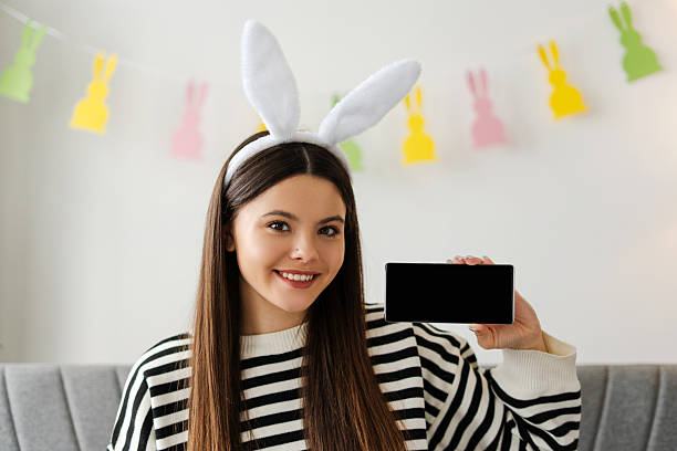 Young girl wearing bunny ears headband smiling and showing smartphone with blank screen sitting on sofa with easter decorations hanging in the background celebrating. Online technology concept