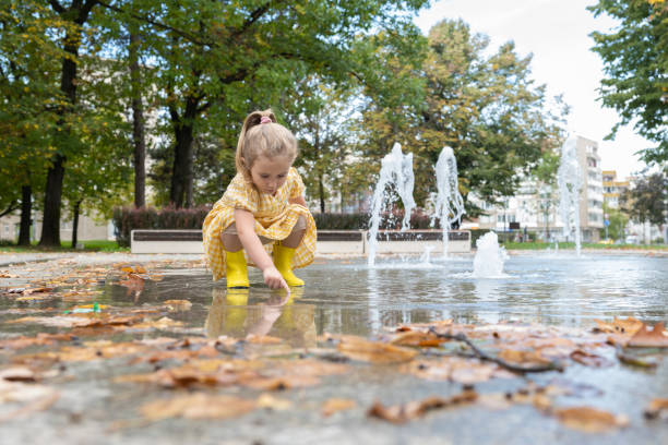 Summer water fun. Young girl in yellow boots is enjoying the summer by the fountains in the city park, her feet splashing in the water.