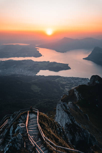 View down fenced pathway to mountains and lakes under sunrise, Mount Pilatus viewpoint above Lake Lucerne, Nidwalden Canton, Swiss Alps