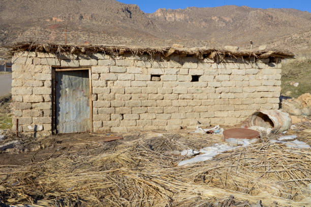 An old mud-brick house stands abandoned in a vast, dry landscape, its cracked walls revealing years of erosion and neglect. The thatched roof, barely holding together, hints at a time when life once thrived here. The ground surrounding it is barren, with scattered straw and stones, emphasizing the harshness of the environment. A single door and two small, dark windows stare blankly, as if guarding the silence of its past. This place tells a story of resilience, solitude, and the inevitable passage of time.