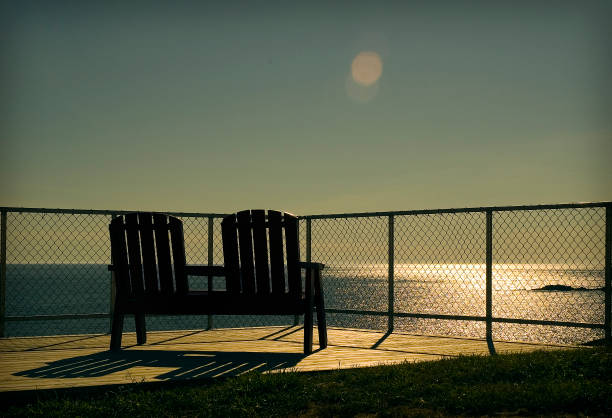 A couple of chairs on a raised vantage point behind a fence over looking the ocean on a clear end of day.