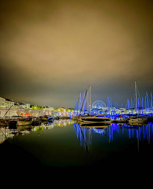 A mesmerizing nighttime view of a marina where sailboats and yachts are docked under a softly lit sky. The still water creates stunning reflections of the boats, harbor lights, and a brightly illuminated Ferris wheel in the distance, which casts a blue glow over the scene. The surrounding buildings add a charming European coastal ambiance with warm lighting contrasting against the cool tones of the night. This image is ideal for themes related to waterfront nightlife, luxury travel, serene coastal settings, and illuminated cityscapes.